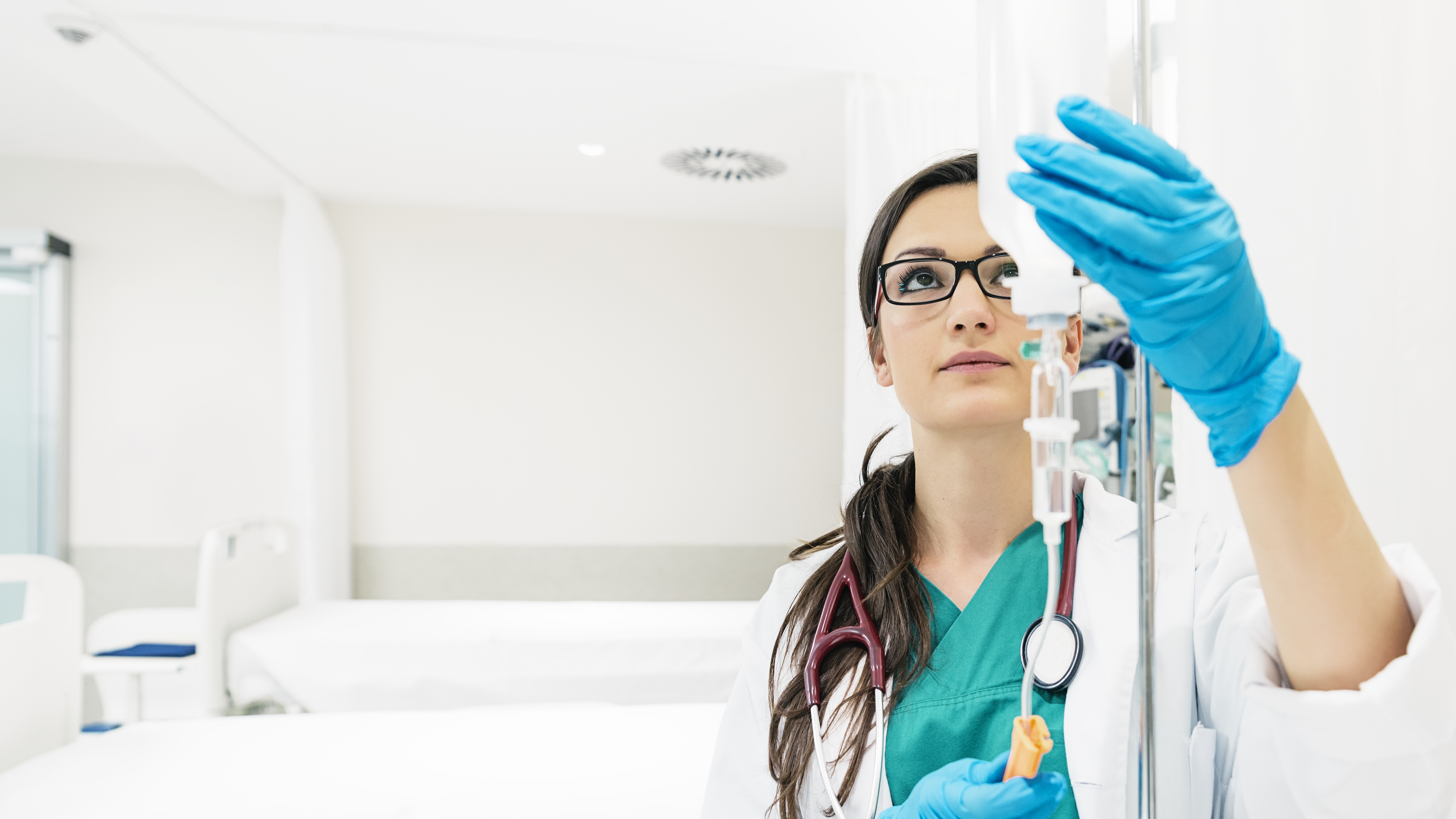 A woman in medical protective clothing with blue gloves touches an IV drip, a hospital room can be seen in the background 